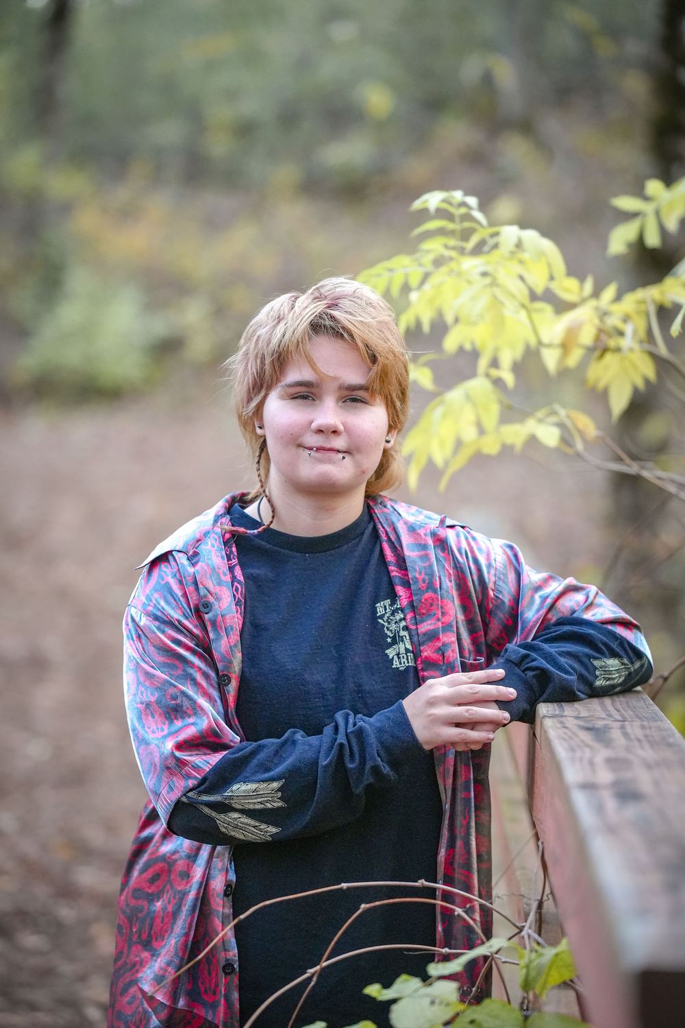 A person in a dark shirt and floral jacket poses outdoors among autumn foliage.