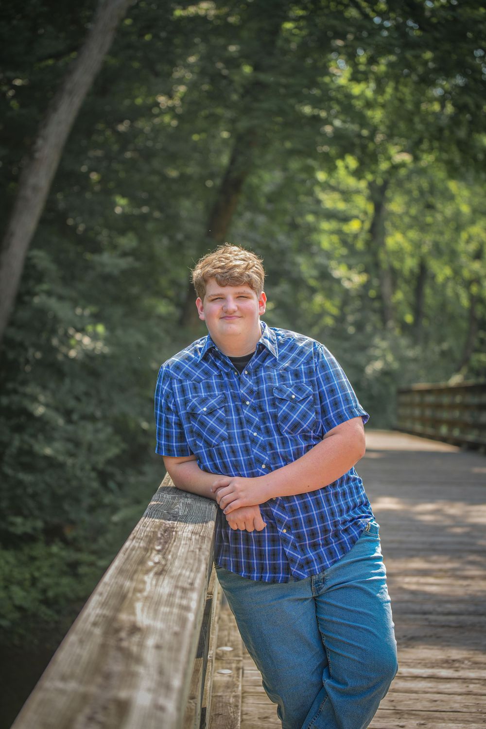 Person in blue plaid shirt and jeans poses on wooden bridge railing in wooded outdoor setting.