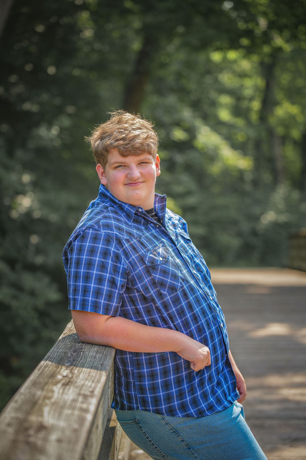 Person in blue plaid shirt and jeans sitting on wooden bench in wooded outdoor setting.