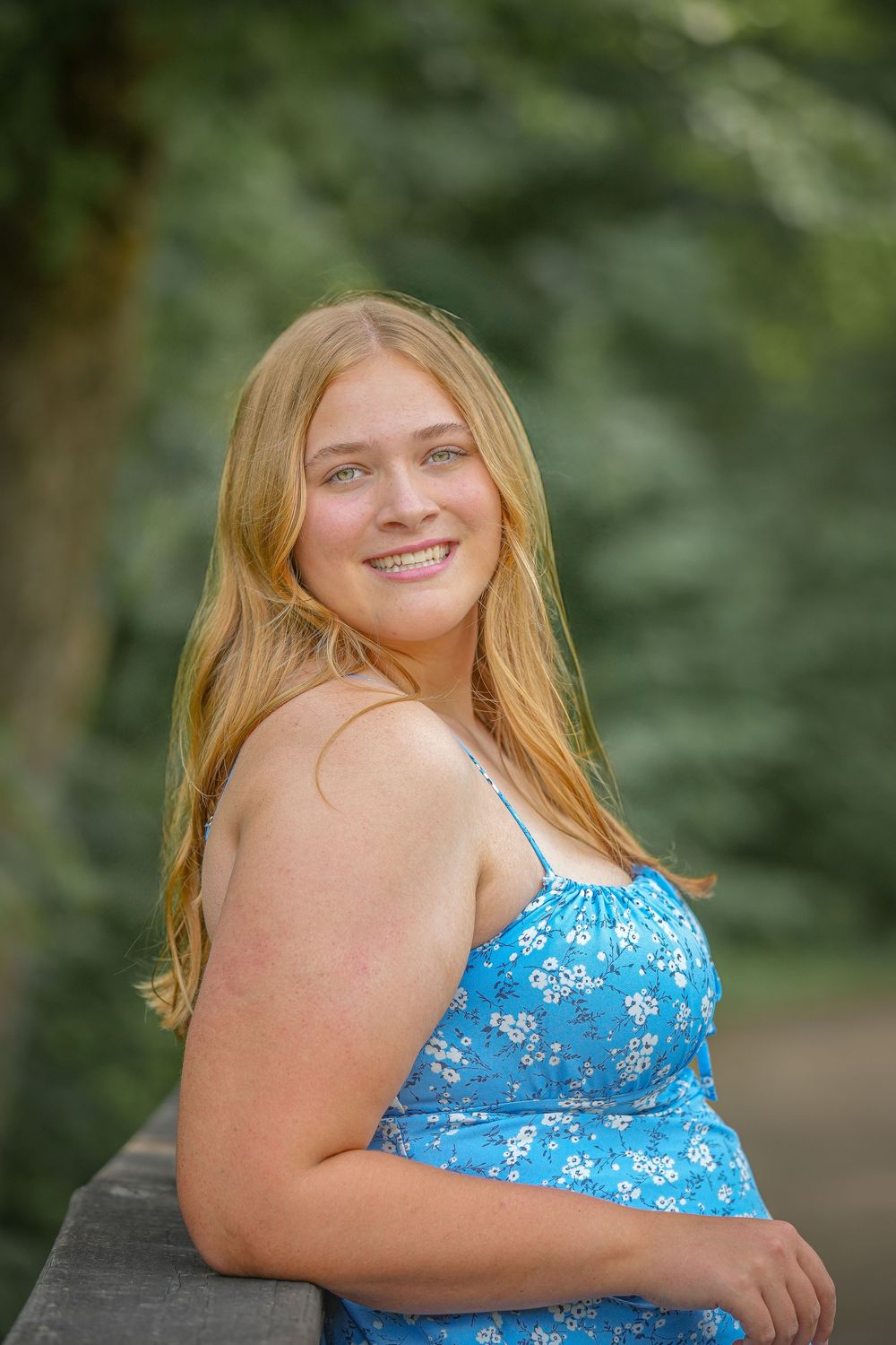 Young woman in a blue floral dress poses outdoors with a natural smile against a blurred forest background.