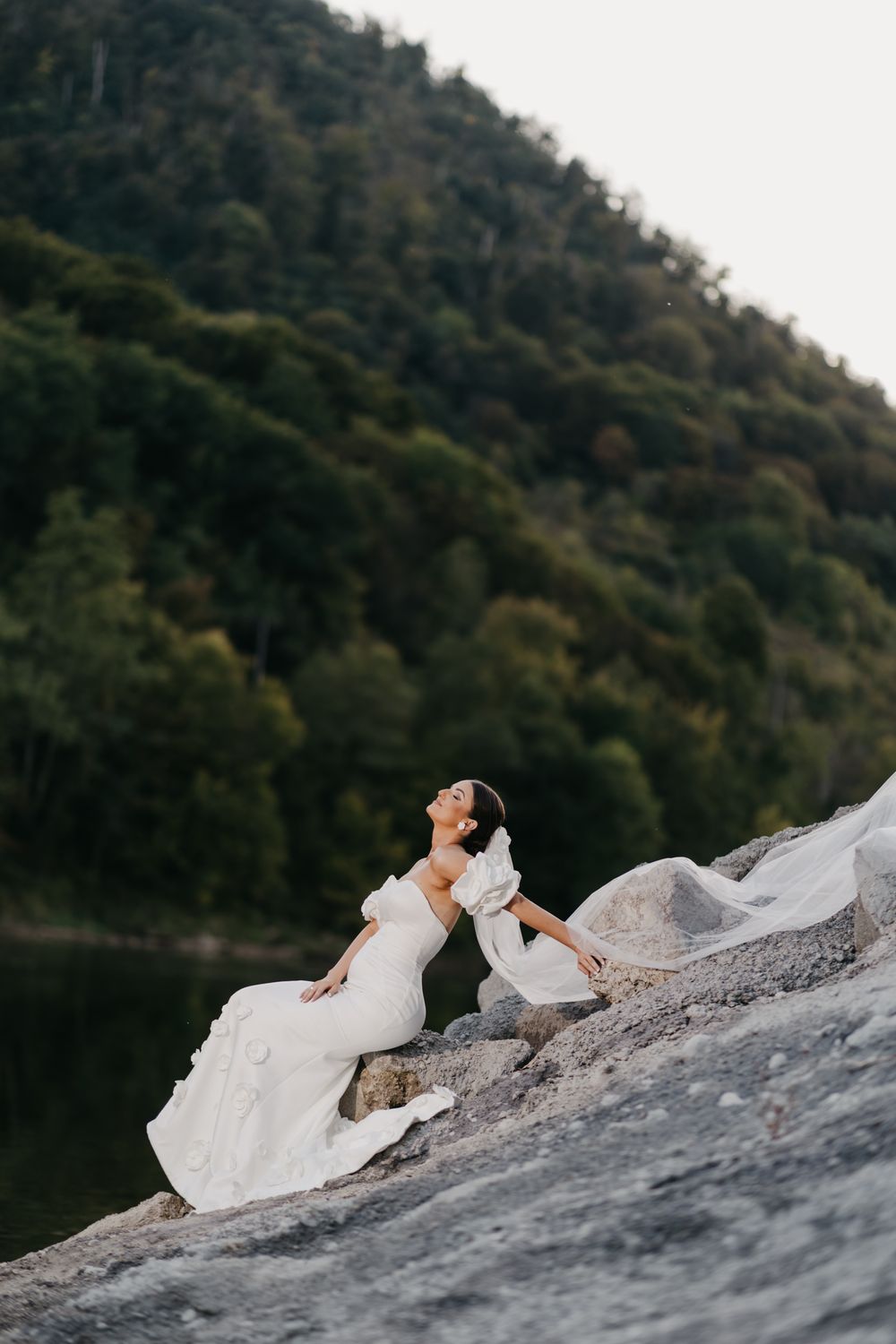A person in a flowing white wedding dress lies gracefully on a rocky surface against a mountainous backdrop.