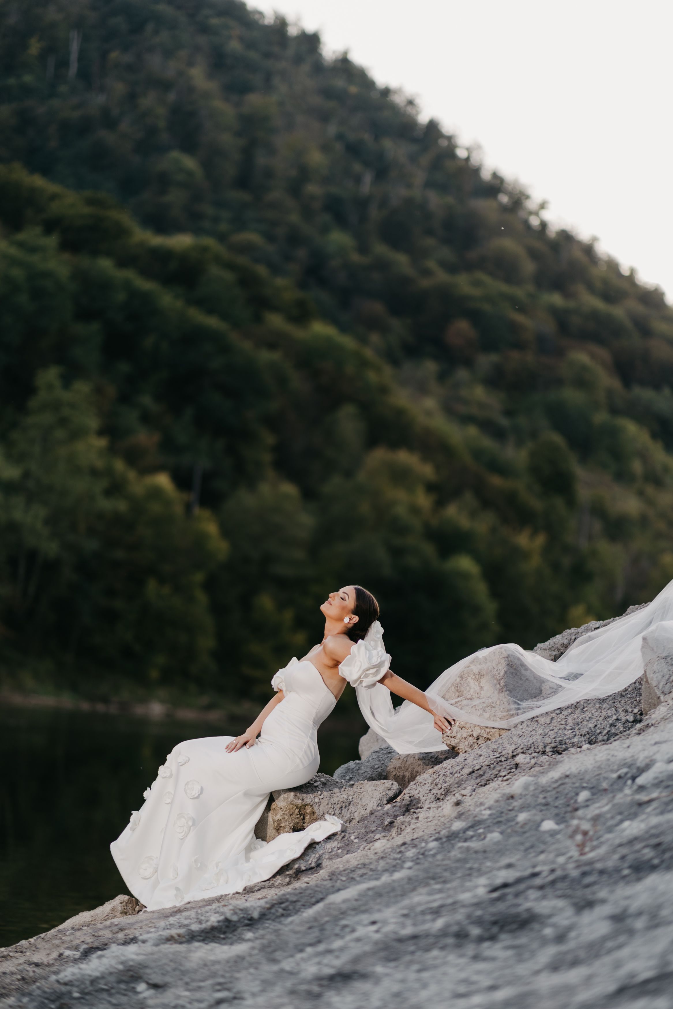 A person in a flowing white wedding dress lies gracefully on a rocky surface against a mountainous backdrop.