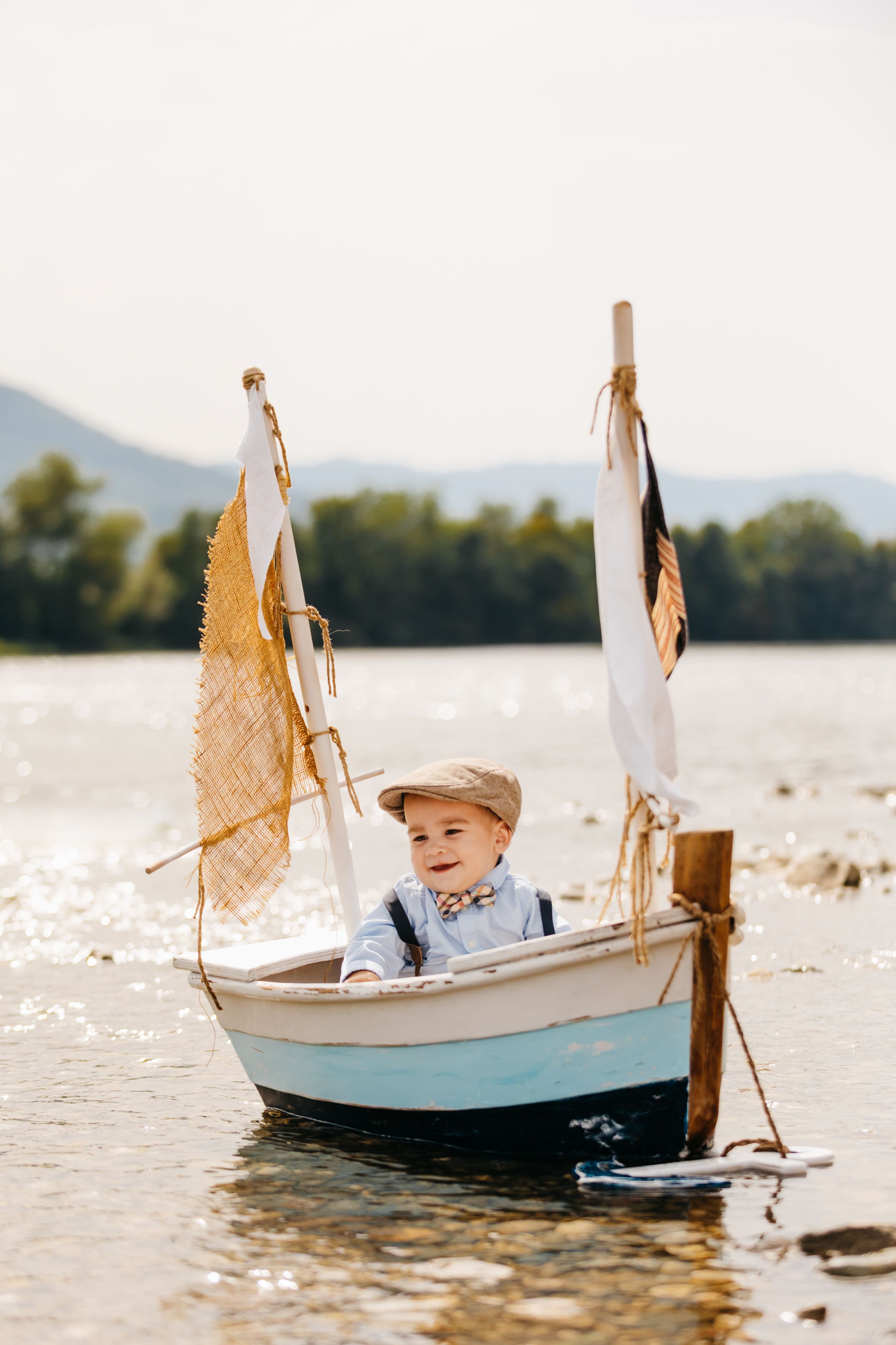 A small white and blue toy sailboat with canvas sails sits in shallow water against a mountain backdrop.
