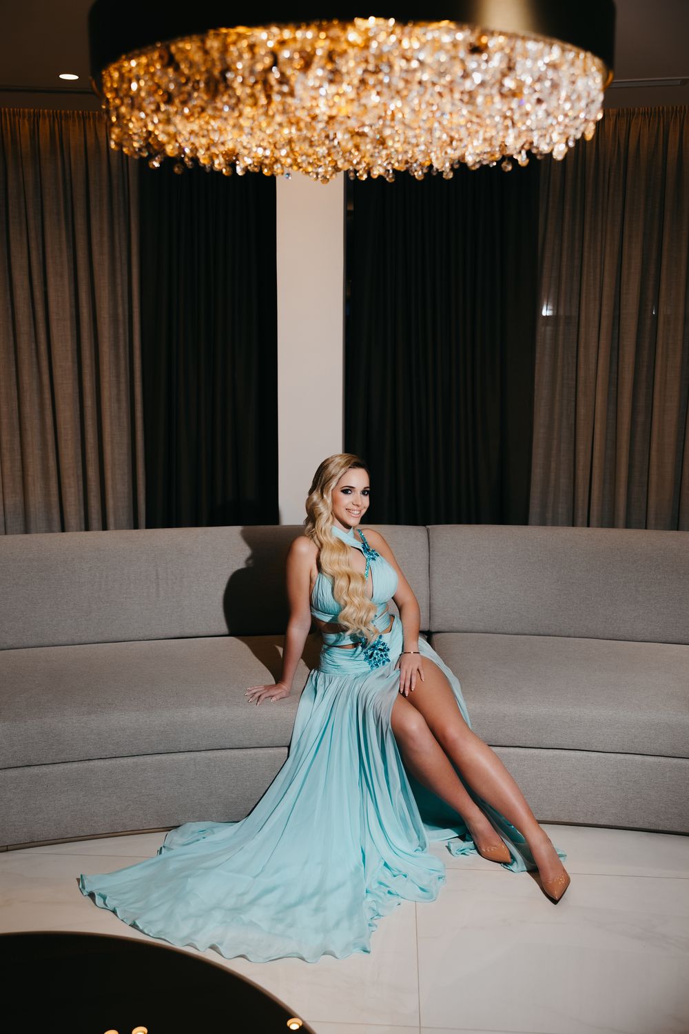 A person in a light blue evening gown poses elegantly on a curved sofa beneath a crystal chandelier.