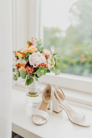 Elegant bridal bouquet with peach and coral flowers next to white wedding shoes on windowsill.