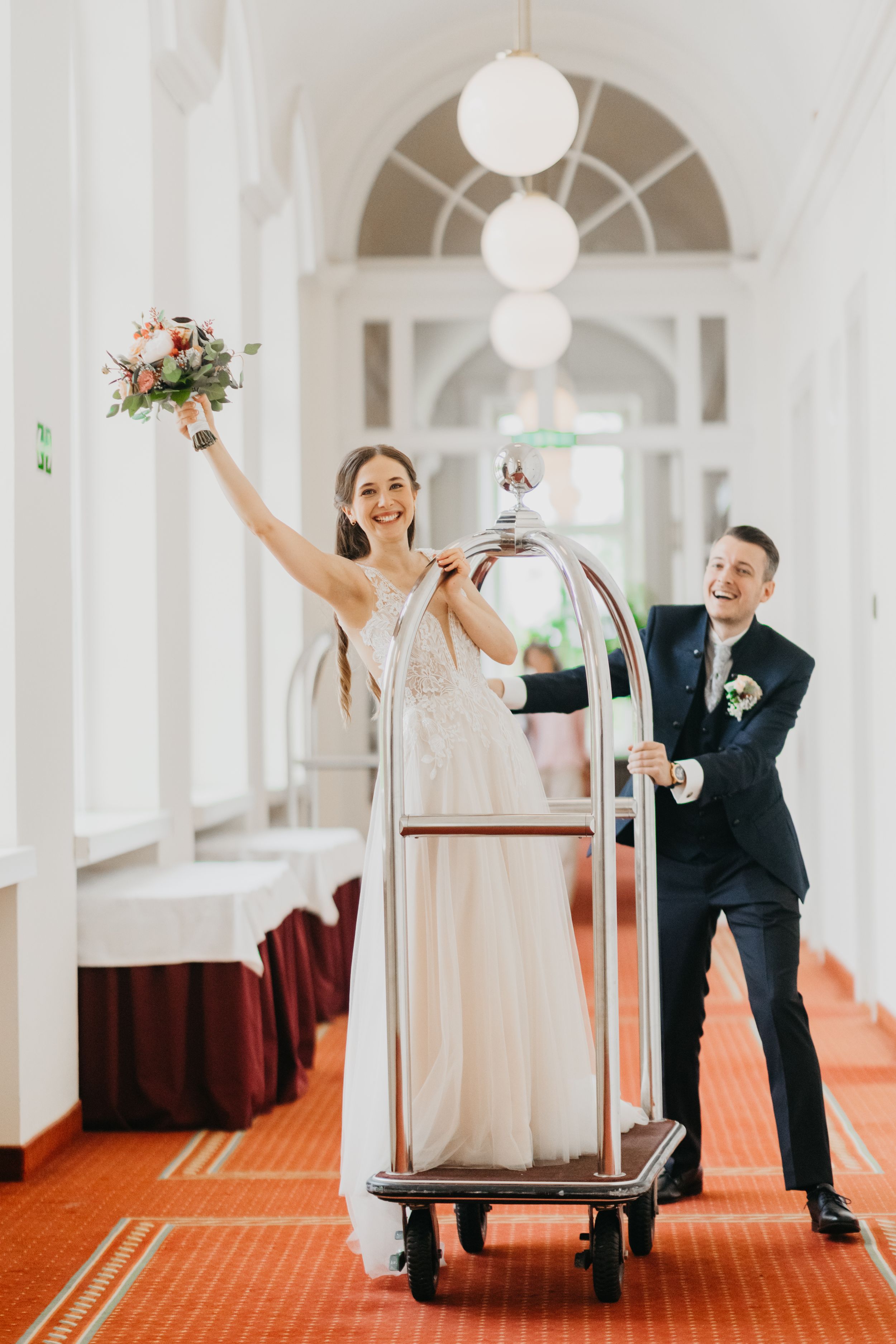 Playful wedding moment with luggage cart in elegant hallway with arched windows.