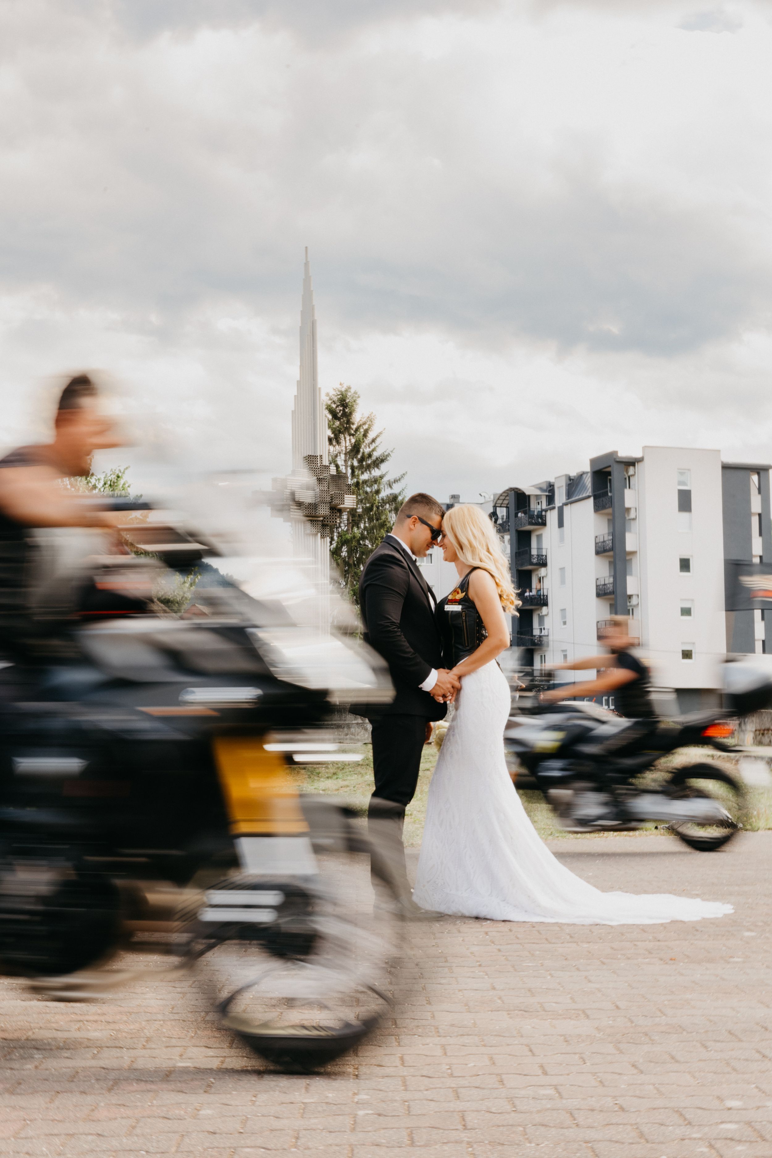 A wedding couple embraces on a street as motorcycles blur past in motion, with urban buildings and church spire in background.