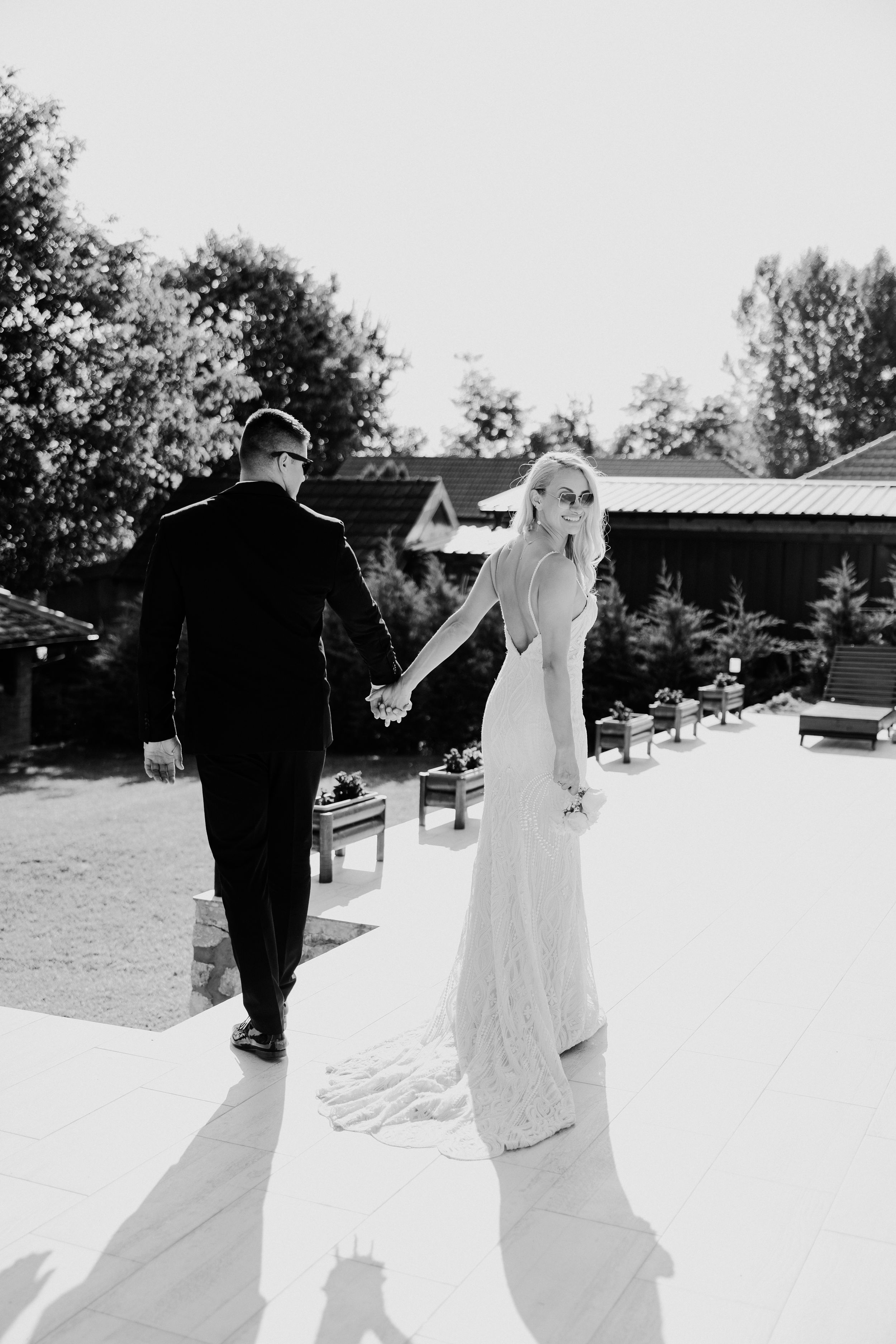 A couple in wedding attire walk hand in hand on a sunny patio surrounded by greenery.