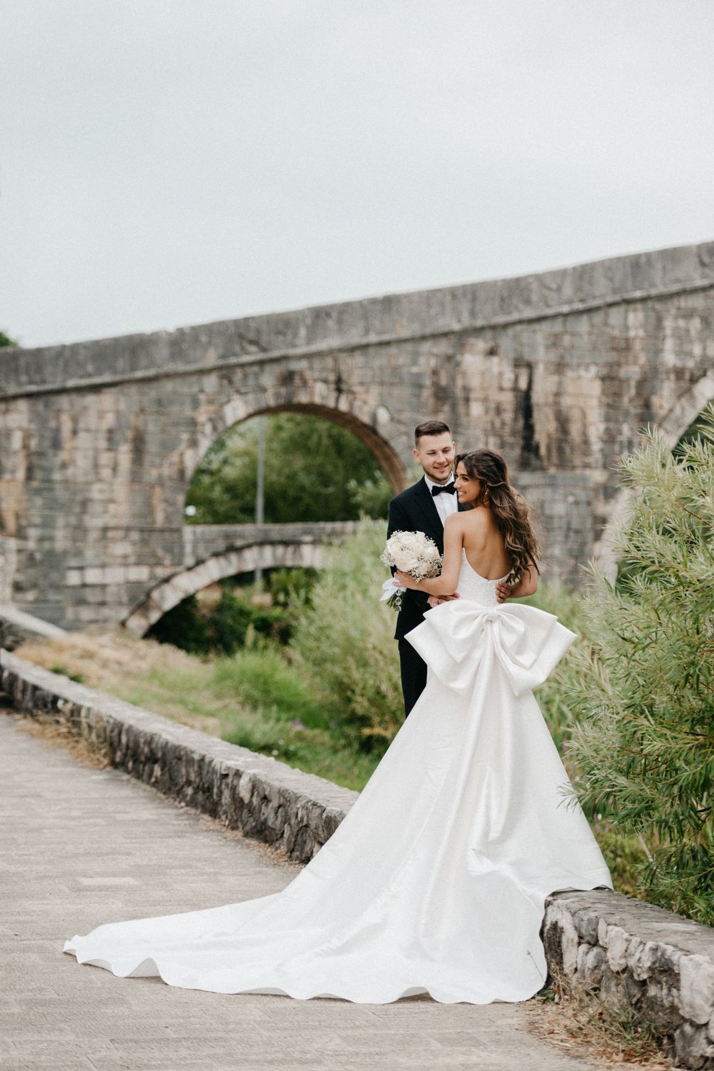 A couple in formal wedding attire embraces on a stone path next to an ancient stone wall.