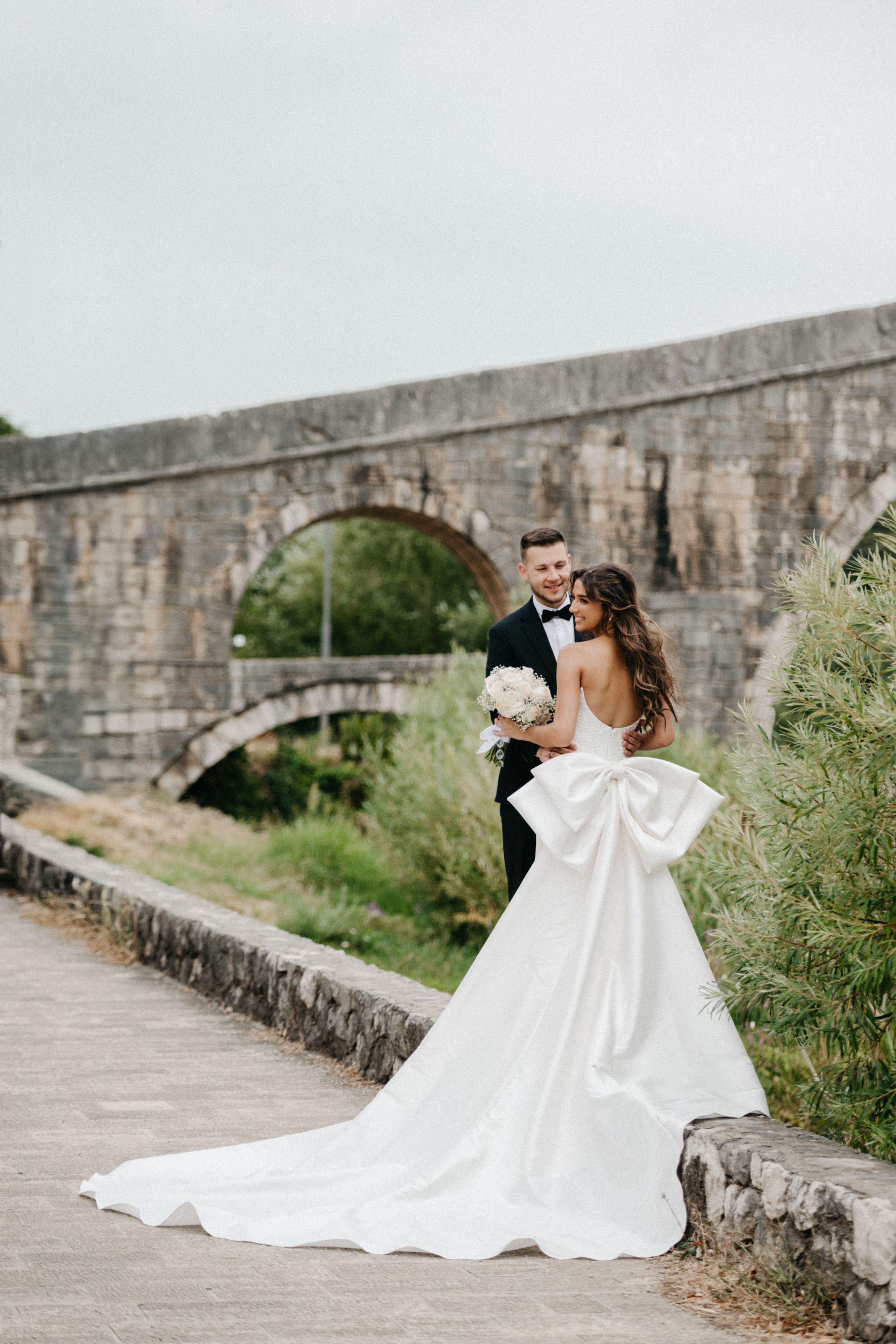 A couple in formal wedding attire embraces on a stone path next to an ancient stone wall.