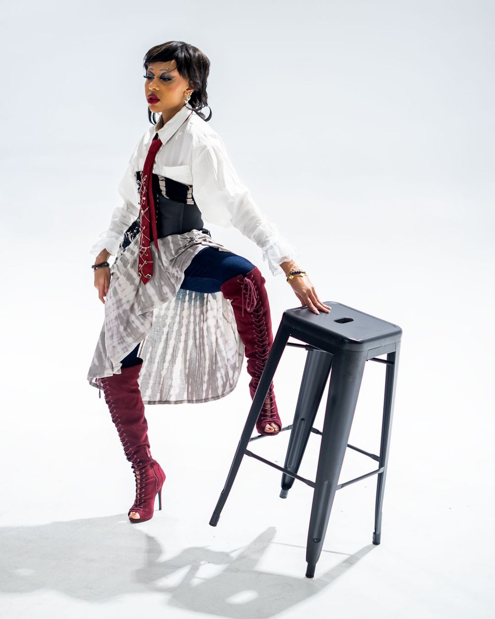 Model in steampunk-inspired outfit with white blouse, corset, burgundy boots and silver skirt poses with black metal stool.
