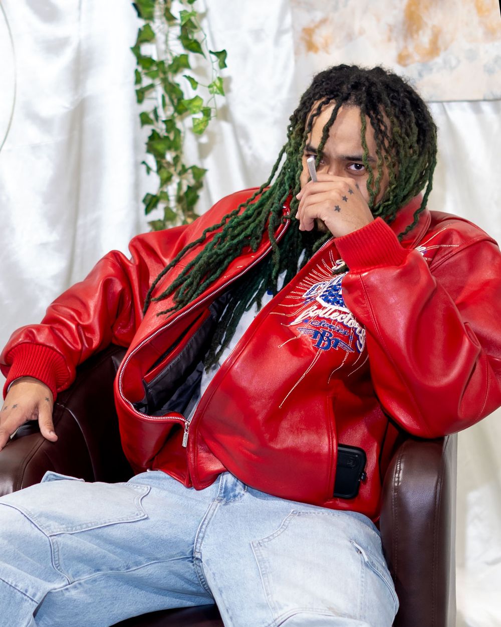 Person in red leather jacket and jeans sitting on brown leather chair with hanging plants in background.
