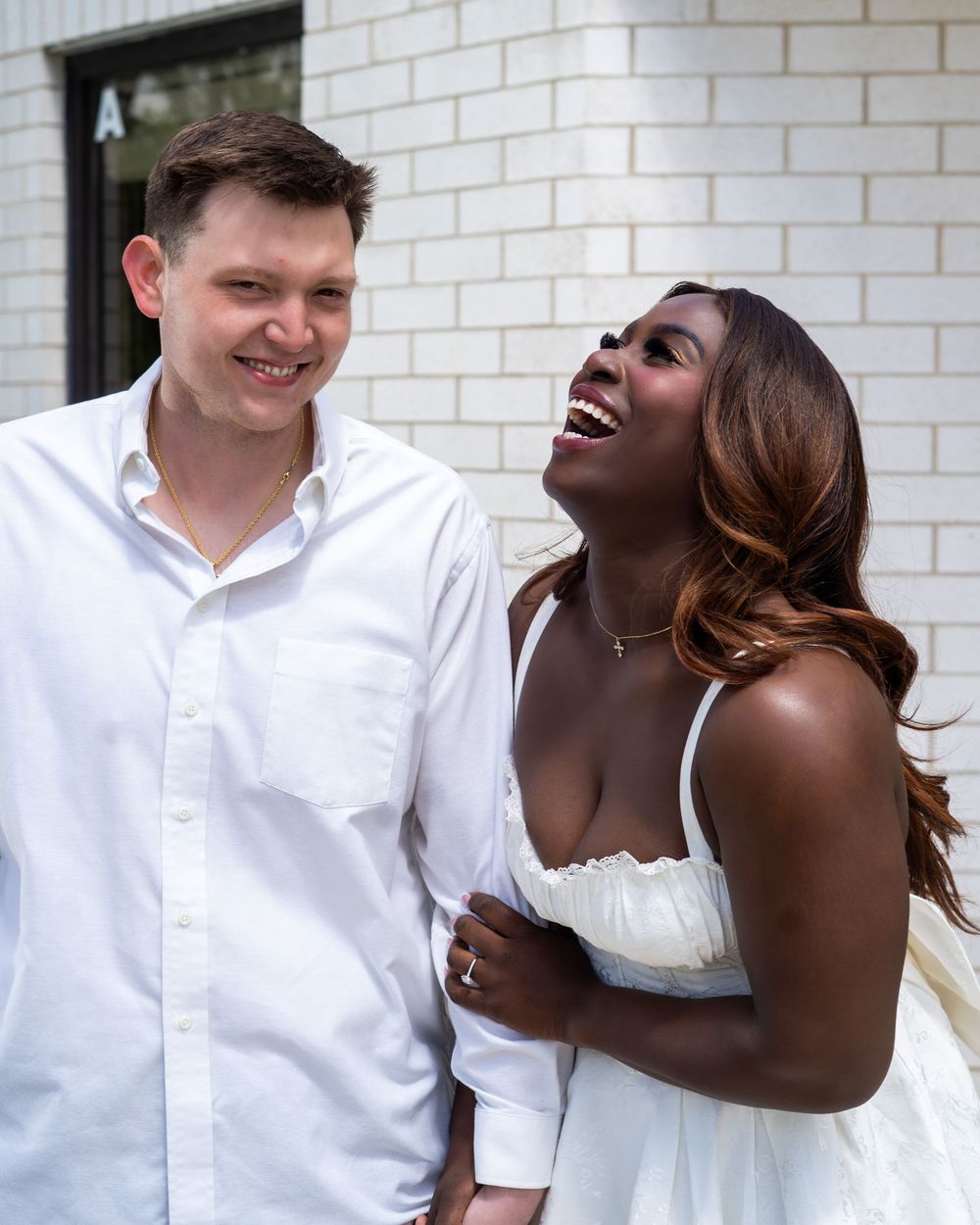A joyful interracial couple laughing together outdoors, dressed in white outfits against a brick wall backdrop.