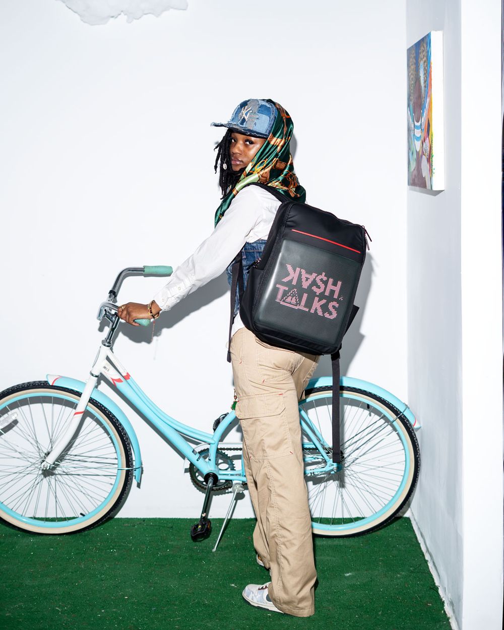 A delivery person with a DoorDash-style backpack stands with a light blue bicycle on artificial turf against a white wall.