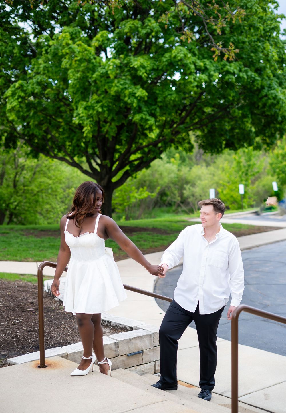 Couple holding hands on steps outdoors, woman in white dress and man in white shirt, lush green trees in background.