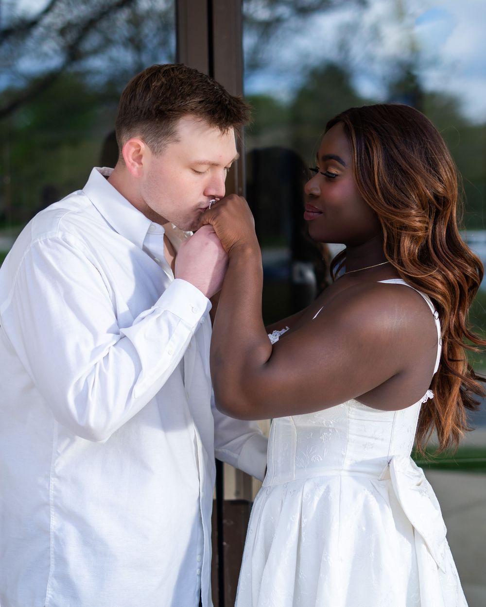 A man in a white shirt kisses a woman's hand during a romantic couples engagement photo session outdoors.