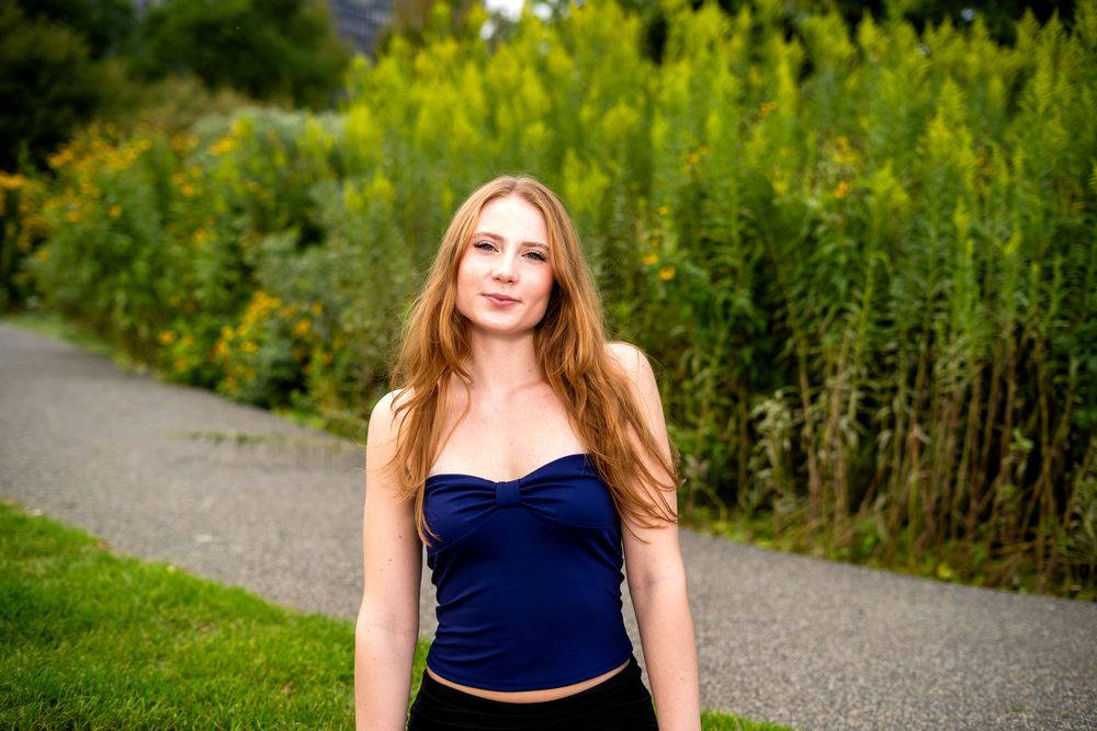 Young woman with long red hair wearing a navy blue crop top, standing on a path surrounded by lush greenery.