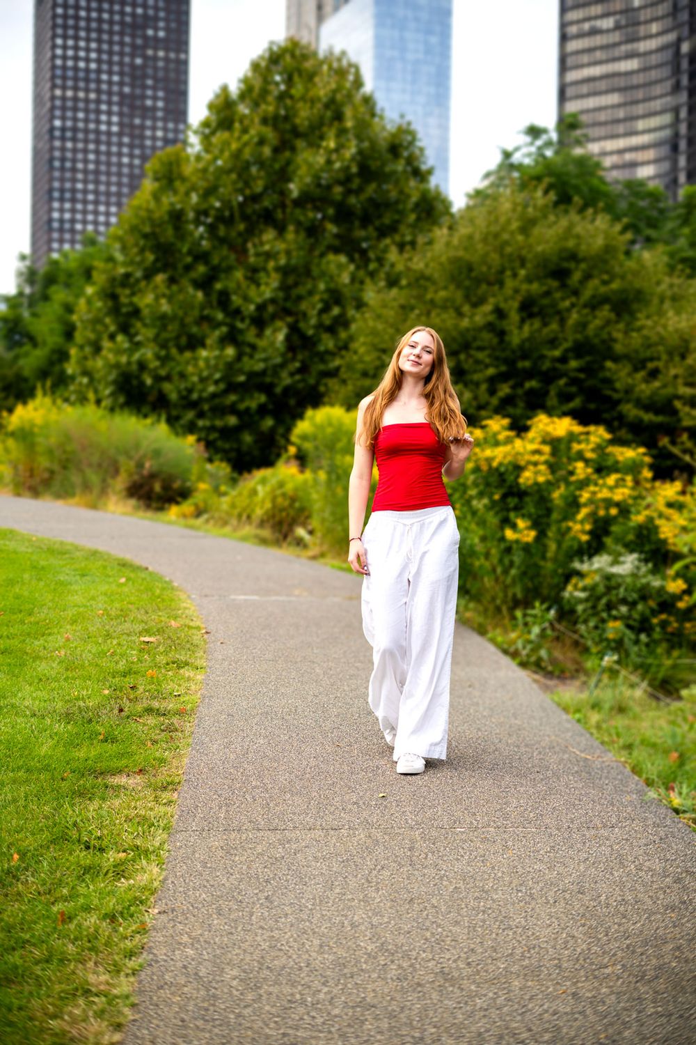 Young woman in red top and white wide-leg pants walking on a park path with city skyscrapers in background.