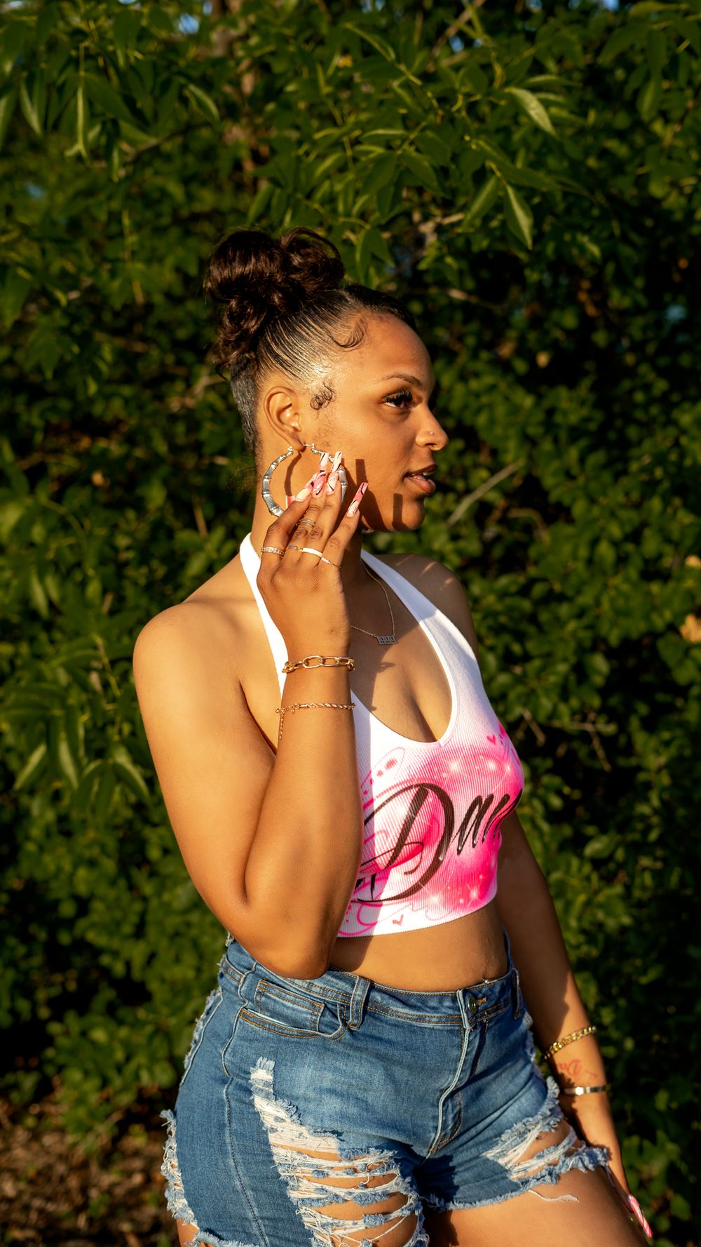 Woman in pink and white halter top poses confidently against green foliage backdrop.