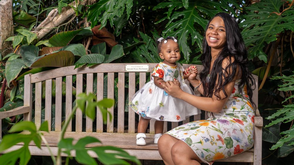 Mother and child in floral dresses share sweet moment on garden bench surrounded by lush greenery.