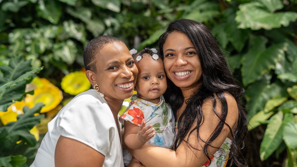Happy family of three pose together outdoors with lush garden and yellow flowers in background.