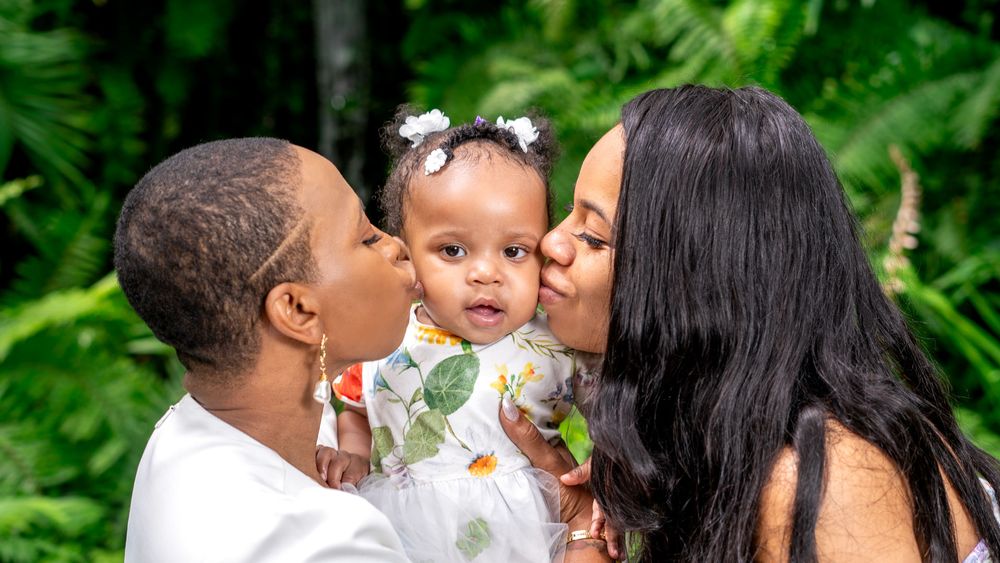 Family sharing tender moment with baby in white dress against tropical garden backdrop.