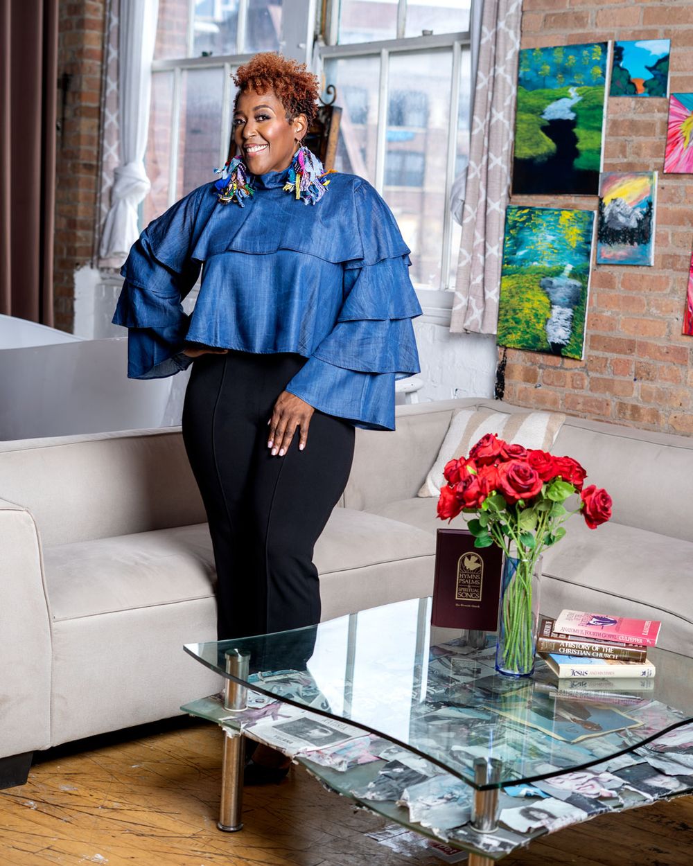 Woman in ruffled denim top and black pants poses in stylish loft with brick walls, art, and red roses.