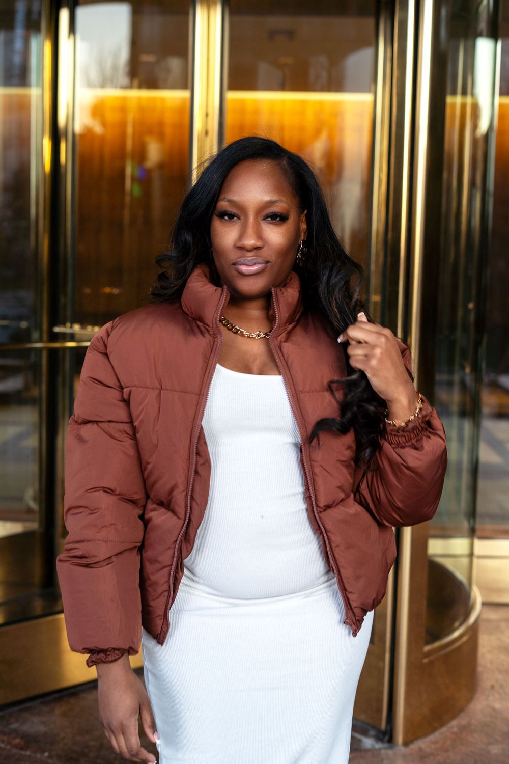 A woman in a brown puffer jacket and white dress poses outside a building with gold revolving doors.