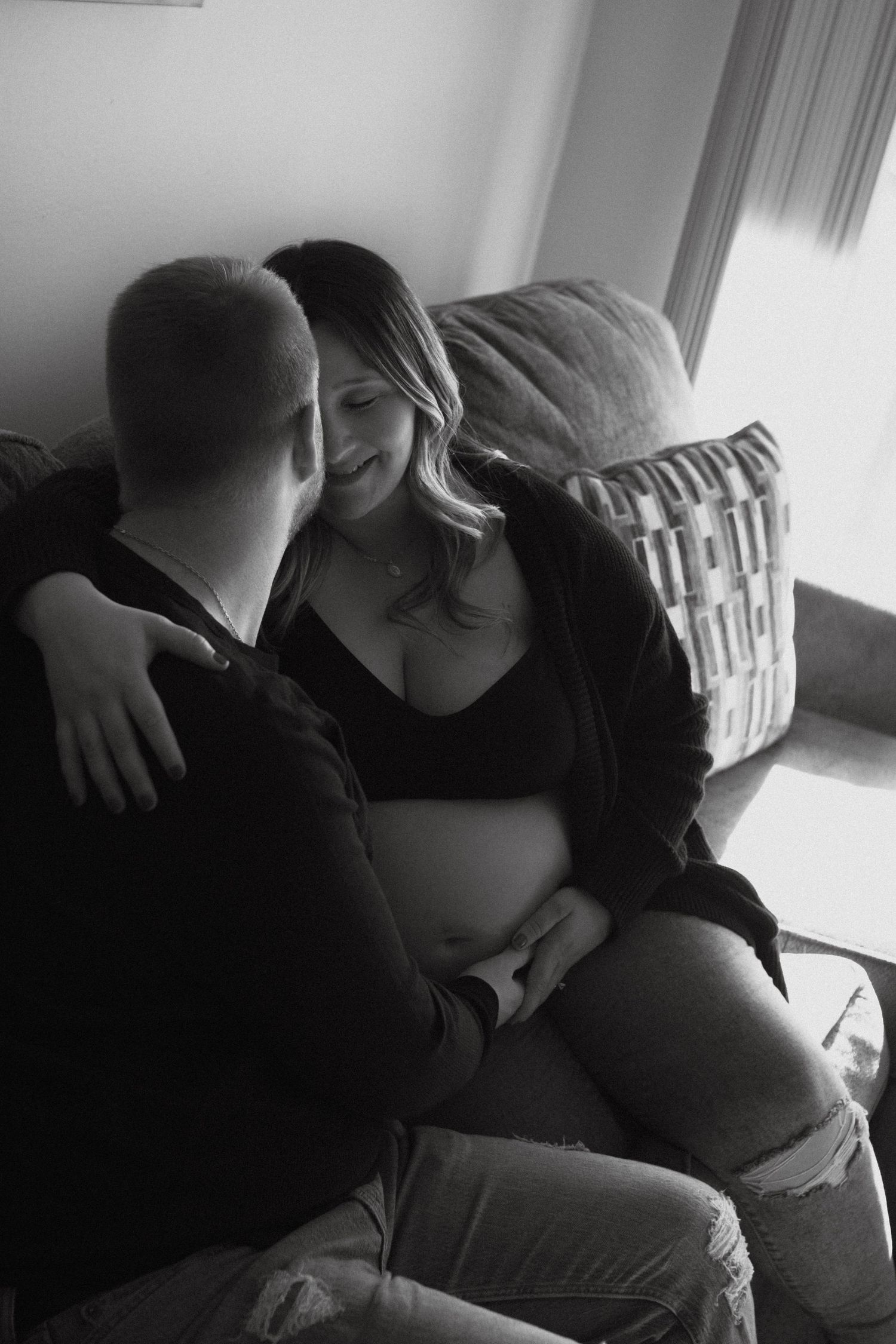 A black and white intimate photo showing a couple embracing on a couch while sharing an affectionate moment together. Maternity photoshoot.