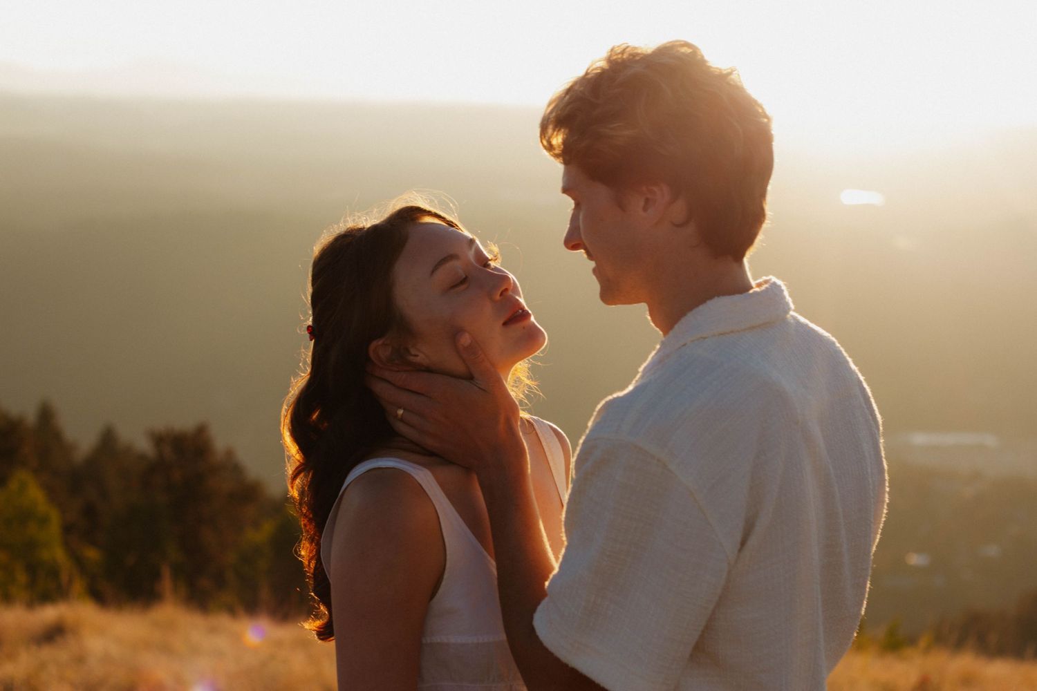 Couple embraces during golden sunset on hilltop overlooking scenic valley view.