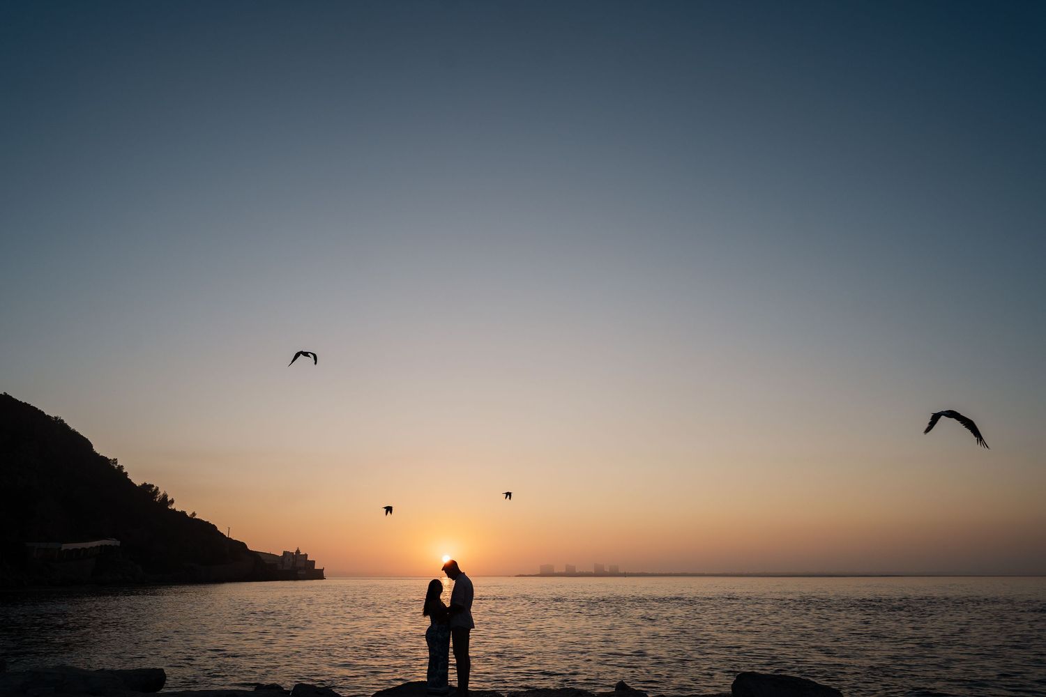 Two silhouettes standing on a grassy hillside against a bright blue sky and horizon.