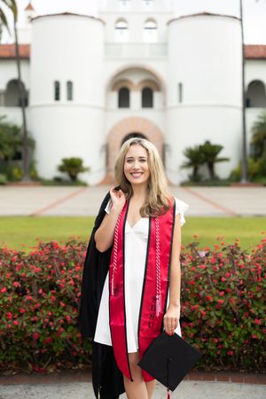 Smiling blonde woman in white dress with red graduation stole and black gown holds cap outside university building.