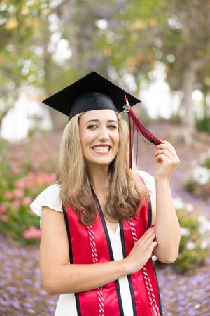 Smiling female graduate in black cap and red sash poses outdoors among colorful flowers for graduation photos.