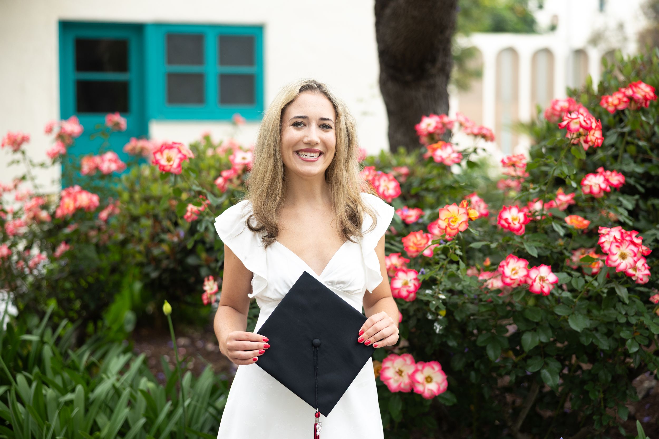 Smiling graduate in white dress holding cap outdoors with pink roses and teal window in background.