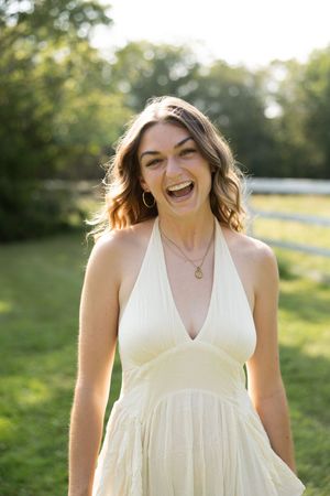A laughing woman in a white halter dress stands outdoors in a sunny green field, wearing gold hoop earrings and a necklace.