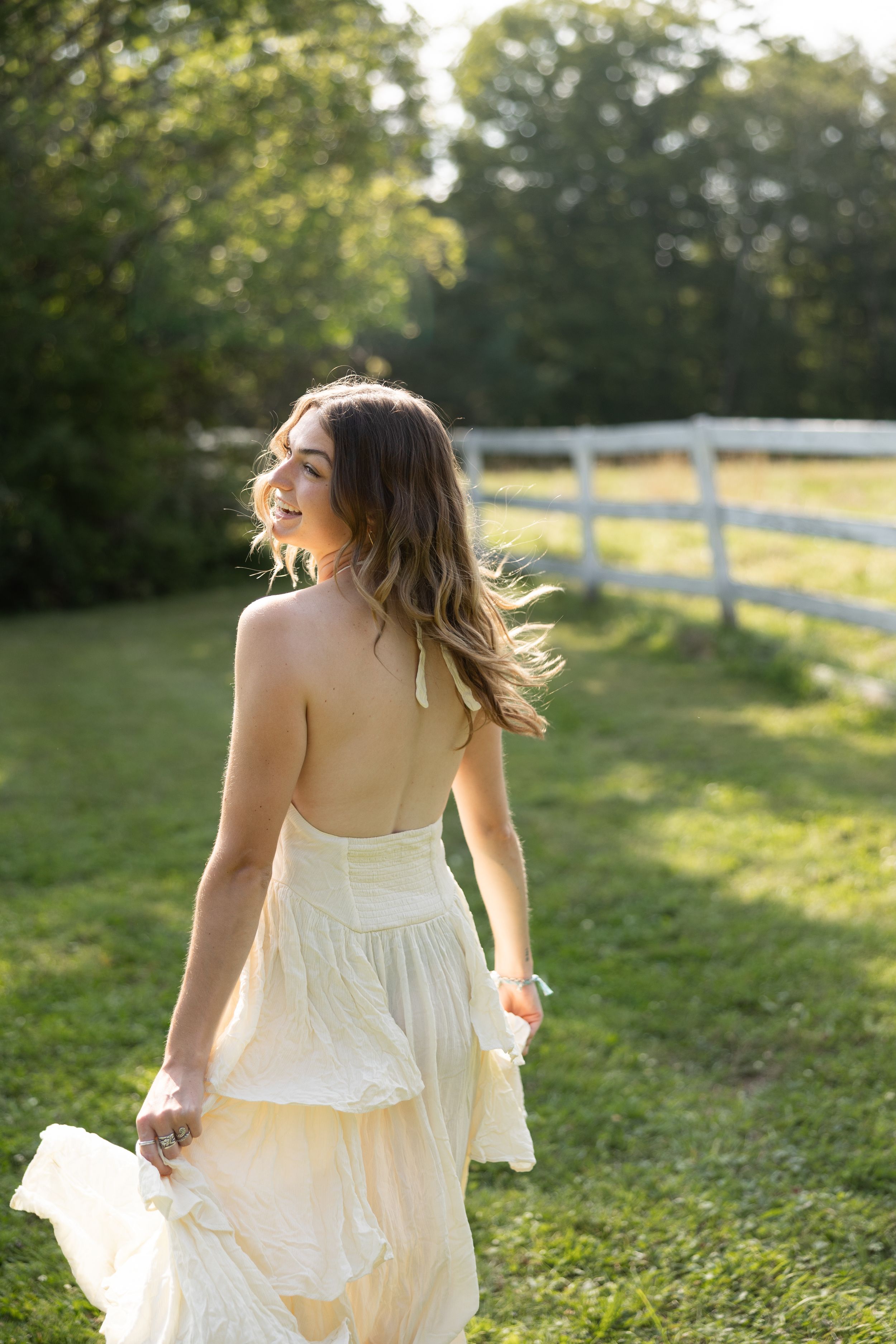 Young woman in white halter dress walking through sunny green field near white fence, hair blowing in breeze.