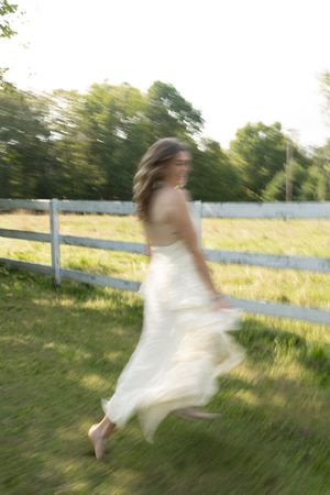 A woman in a flowing white dress runs barefoot through a green field beside a wooden fence on a sunny day.