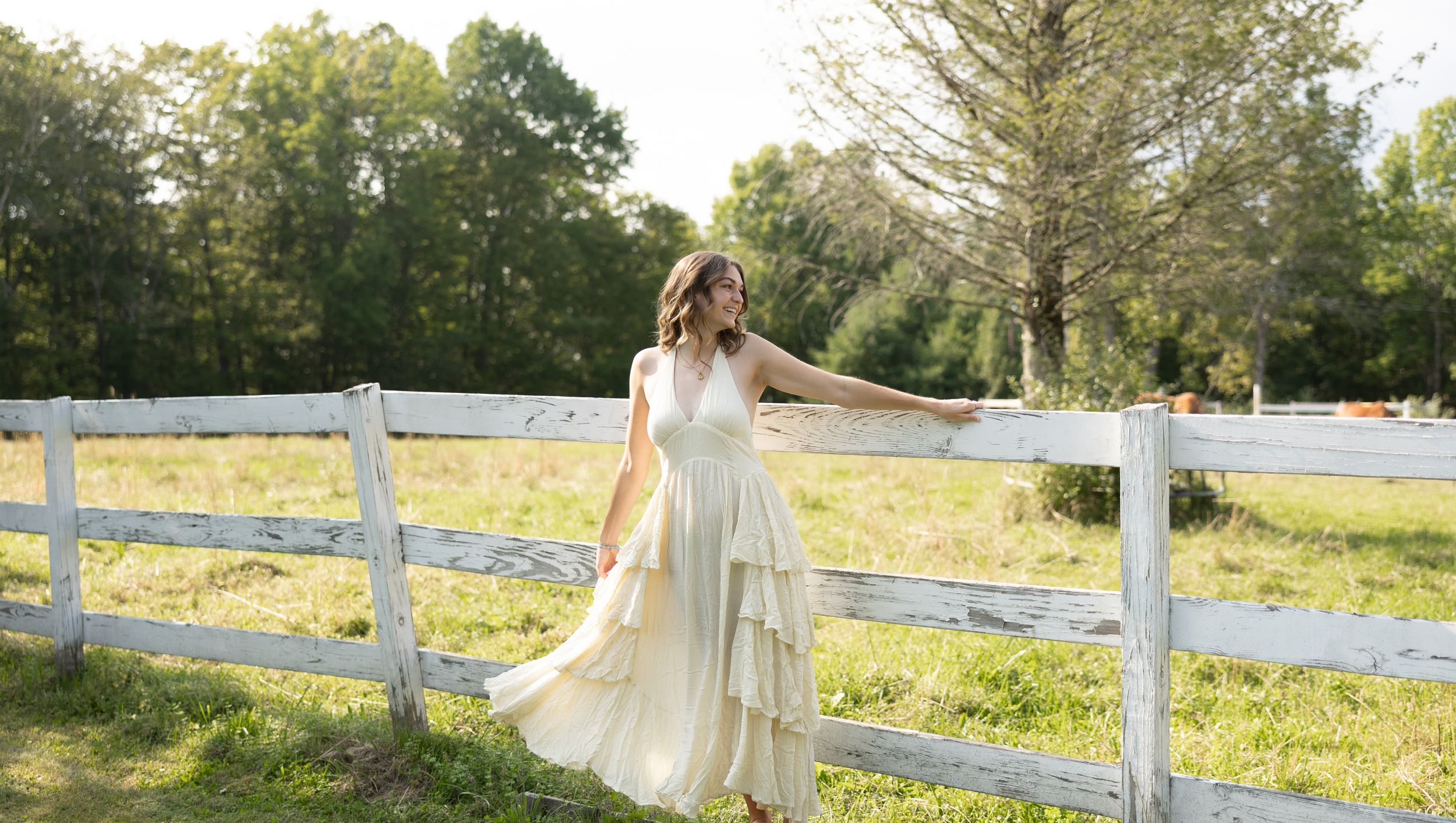 Woman in flowing cream tiered dress poses gracefully by white wooden farm fence with lush green trees in background.