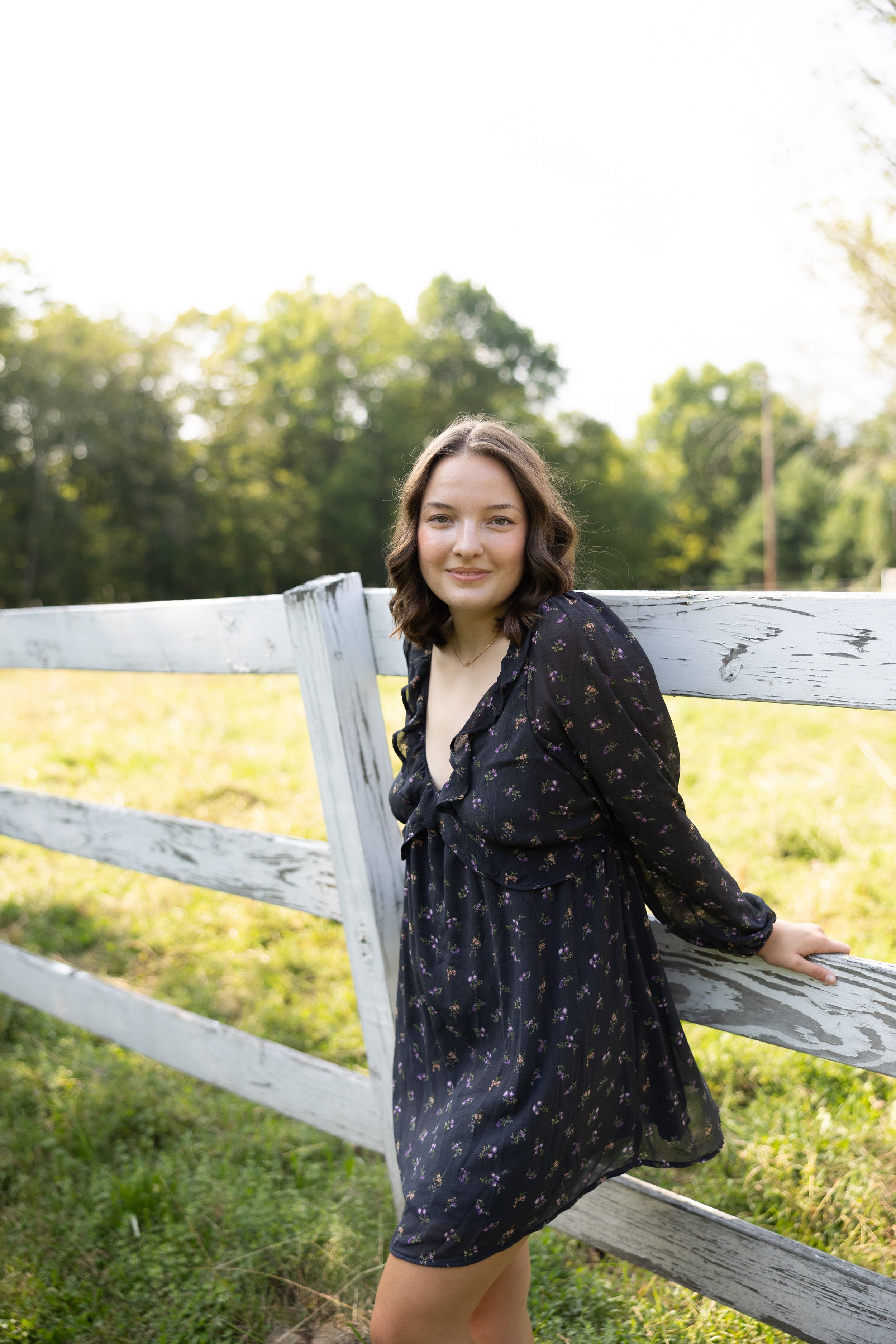 Young woman in black floral dress leaning against white wooden fence in sunny green field.