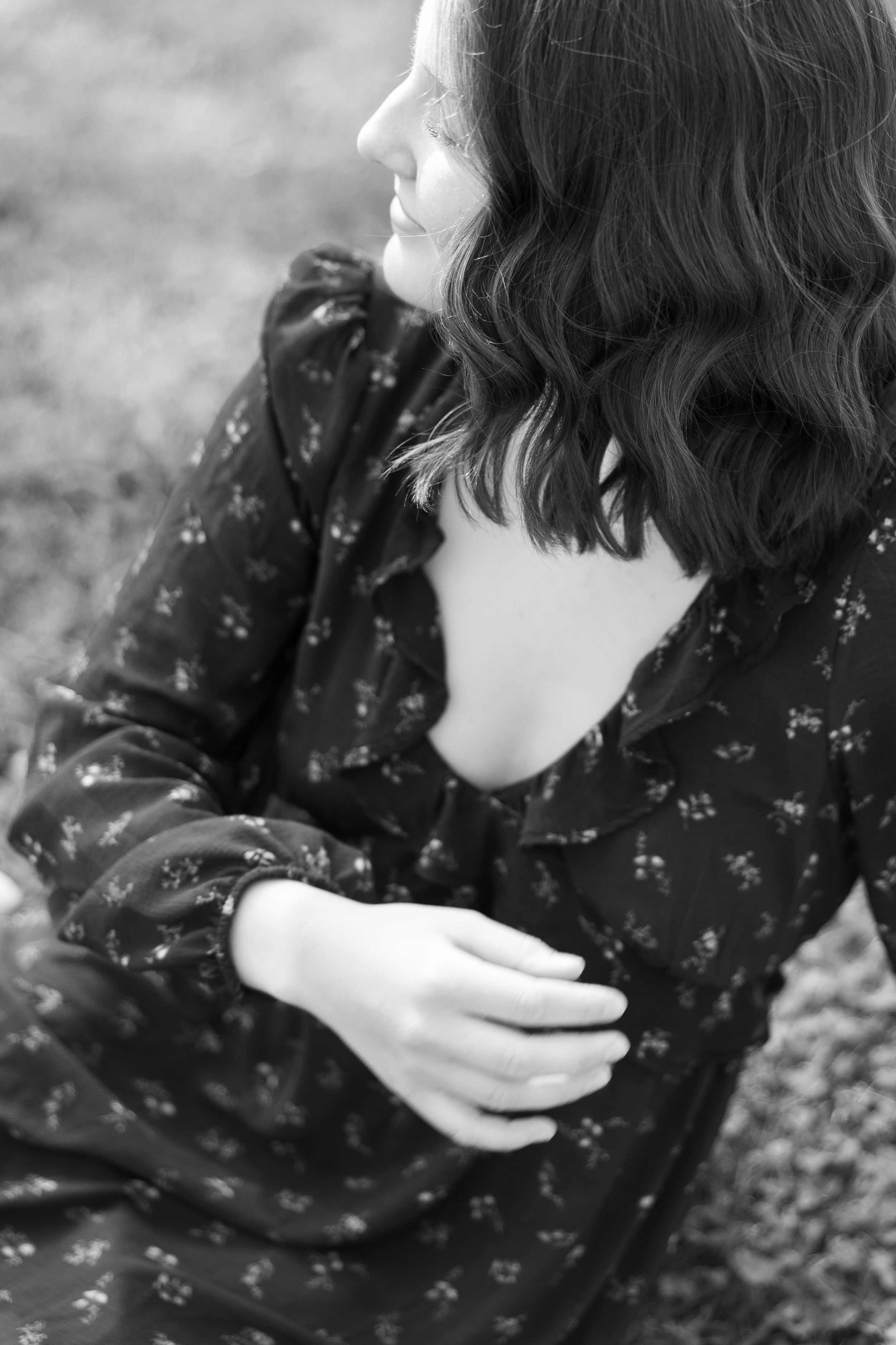 Black and white portrait of a young woman in floral dress, sitting outdoors with hands folded, looking to the side.