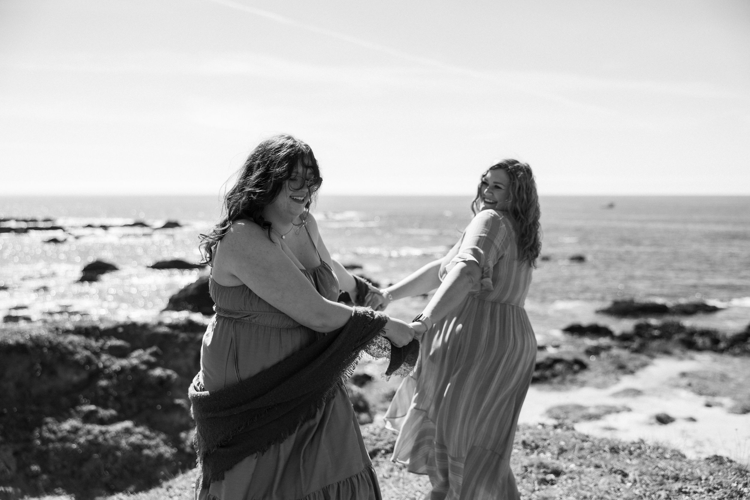 Two women in flowing dresses hold hands and dance near a rocky ocean coastline.