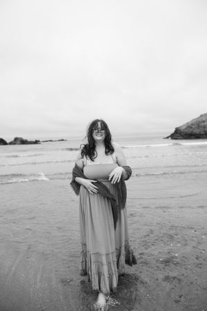 Woman in flowing dress standing on beach shore in black and white photography.