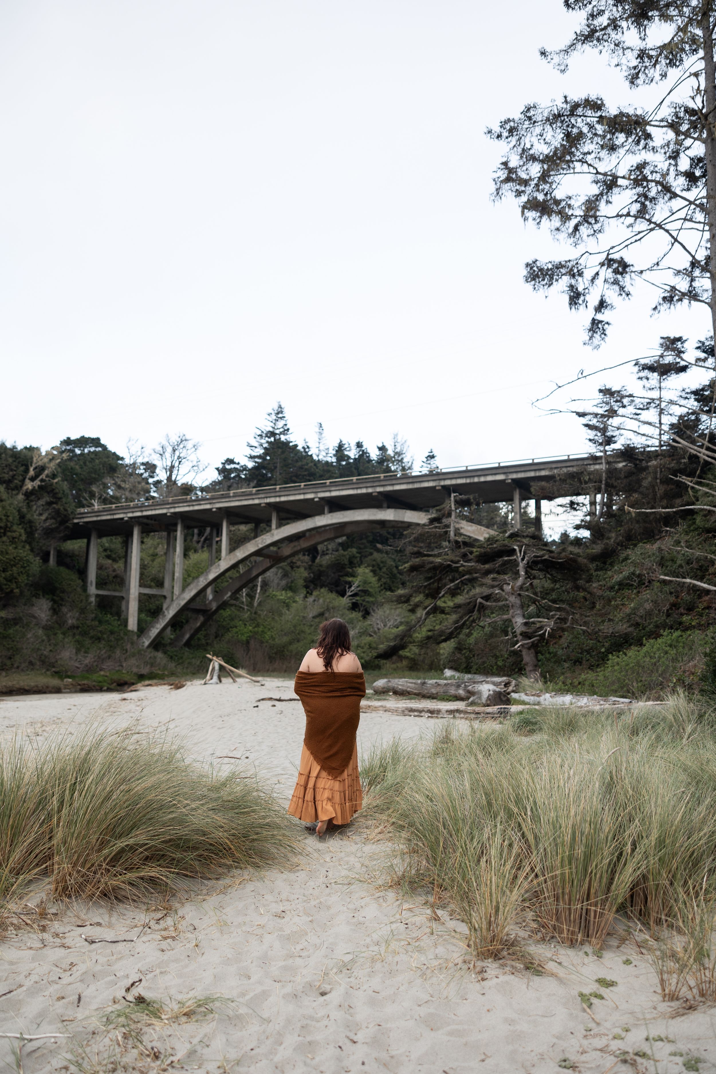 Woman in a brown wrap and skirt walks toward an arched bridge on a sandy beach path.