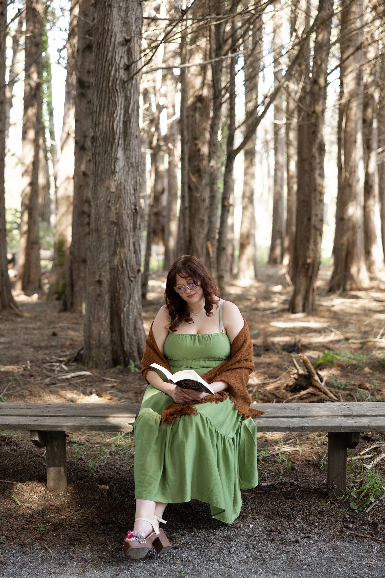 Woman in green dress reading book on bench in sunlit forest setting.