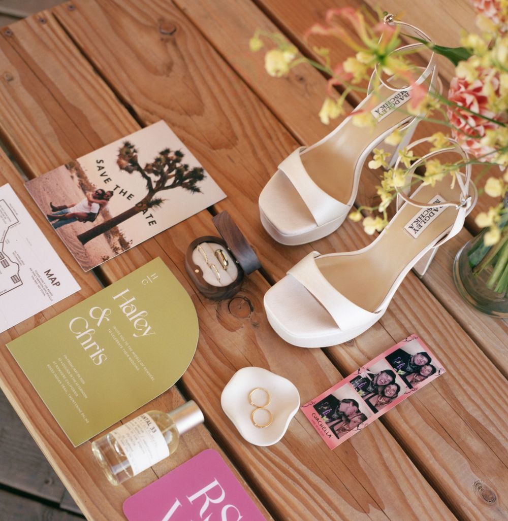 White bridal heels displayed with wedding invitations, rings, perfume, and floral accents on a wooden surface.