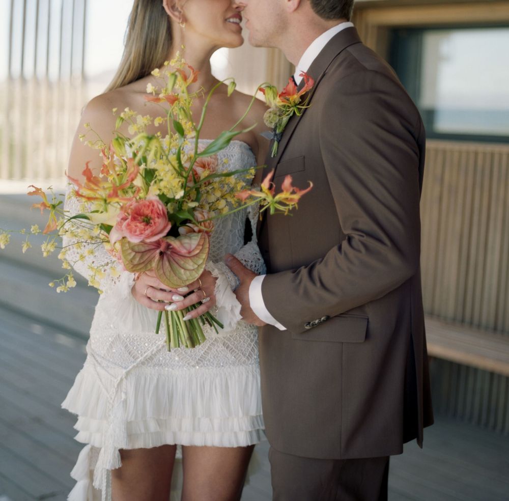 Bride in white mini dress holds colorful bouquet while kissing groom in brown suit outdoors.