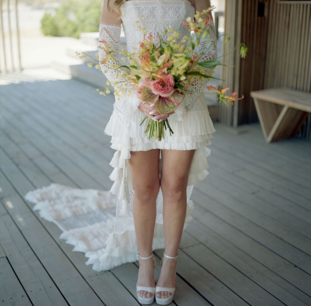 Bride in short white lace dress with train holds colorful spring bouquet on wooden deck.