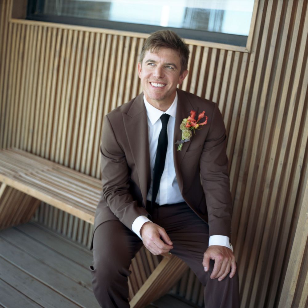 Smiling groom in brown suit with floral boutonniere sitting on wooden bench at outdoor wedding venue.