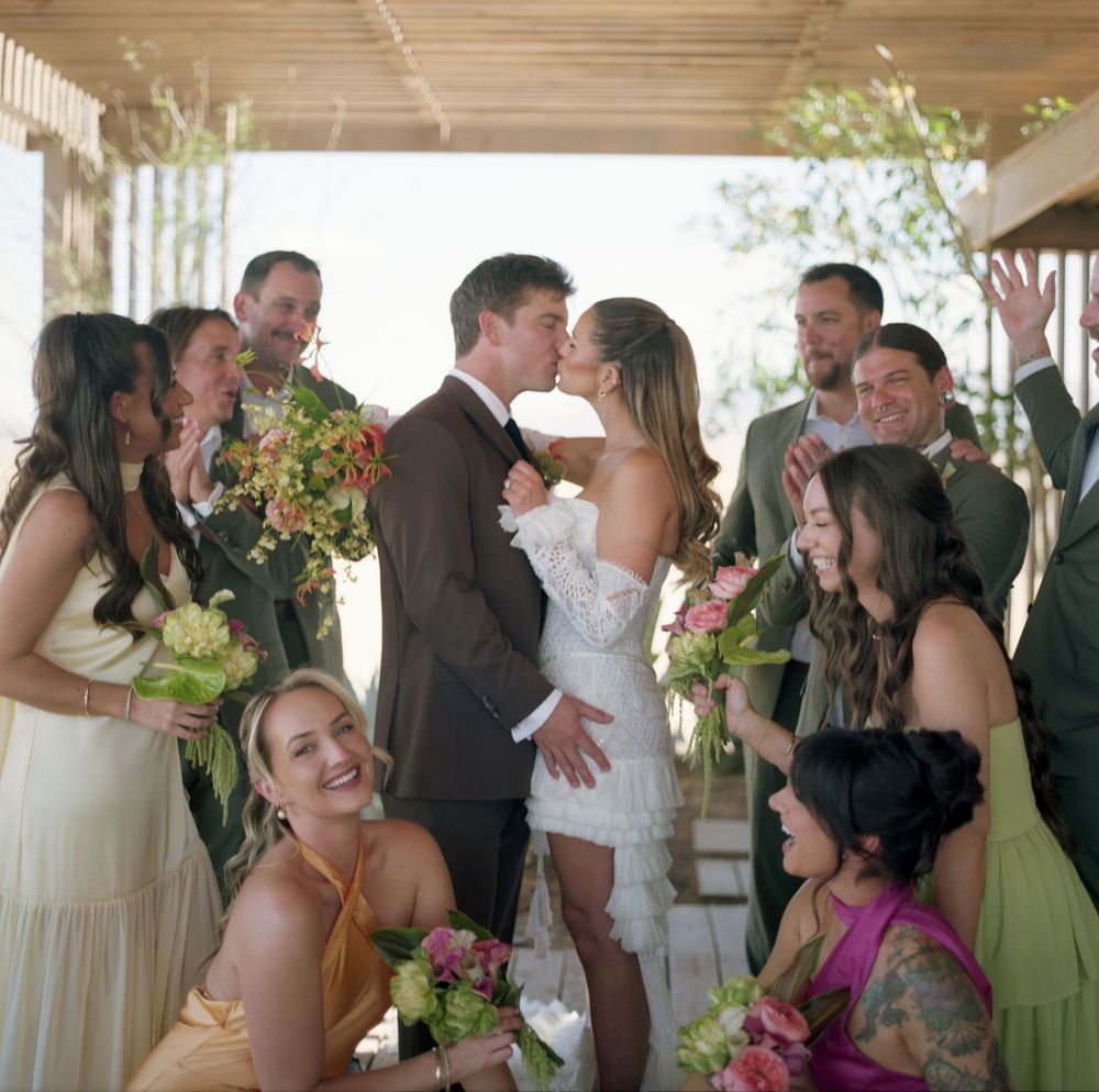 A joyful wedding party surrounds a kissing bride and groom, with bridesmaids holding colorful bouquets outdoors.