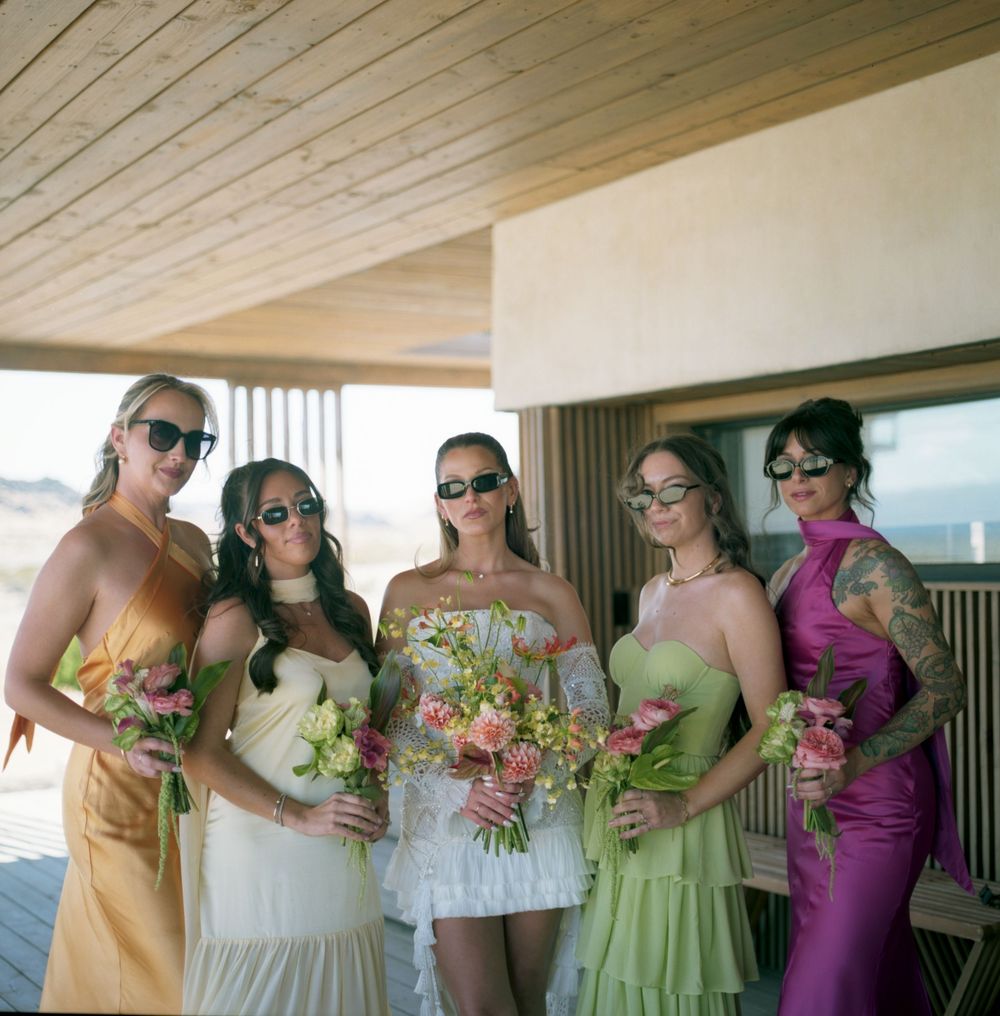 Bride and four bridesmaids in colorful dresses pose with bouquets wearing sunglasses at outdoor venue.